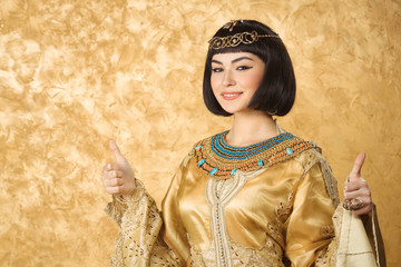 Happy smiling Egyptian woman like Cleopatra with thumbs up gesture, on golden background