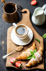 Wall Mural - Breakfast set. Freshly baked croissants with strawberry, mint leaves and cup of coffee on wooden board served with pitcher and copper coffee pot over dark wooden backdrop, top view, selective focus