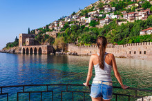 Young Woman Look On Ancient Shipyard Near Of Kizil Kule Tower In Alanya Peninsula, Antalya District, Turkey, Asia. Famous Tourist Destination With High Mountains. Part Of Ancient Old Castle. 