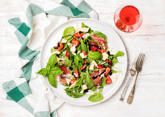 Wall Mural - Summer arugula, prosciutto and strawberry salad with glass of rose wine over white painted wooden background, top view, horizontal composition