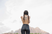 Rear View Of A Topless Young Woman With Long Brown Hair In Braids, Wearing Jeans, Tattoo Of A Celtic Cross On Her Back.