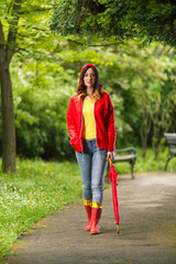 Wall Mural - Portrait of a beautiful young woman in hooded red raincoat looking at camera and smiling while walking in the park on a rainy day.