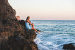 Young blond woman tourist in blue dress sittig on rocks by the sea at sunset. Kleopatra beach, Alanya, Mediterranean region, Turkey.