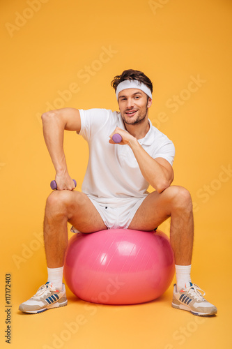 Fotografie, Obraz Smiling sportsman sitting on the fitness ball holding dumbbells