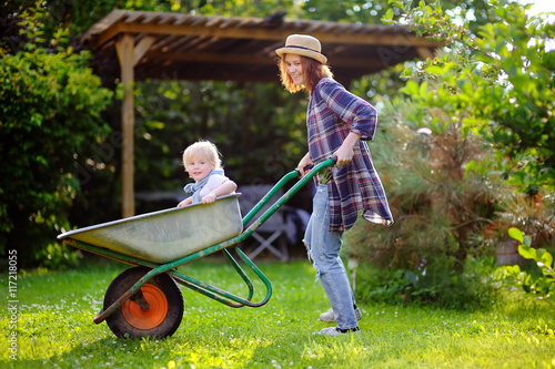Fotografía Adorable toddler boy having fun in a wheelbarrow pushing by mum in domestic gard