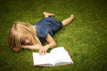Cute Little Blond Girl Reading Book Outside On Grass