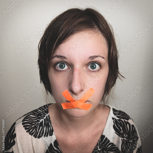 Portrait einer jungen Frau, deren Mund mit Klebeband zugeklebt ist. Sie hat einen erschrockenen Portrait einer jungen Frau, deren Mund mit Klebeband zugeklebt ist. Sie hat einen erschrockenen