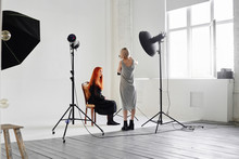 Girl Photographer Photographing Fashion Model In Black Sitting On A Chair On White Background In Studio