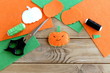 Halloween mini pumpkin decor. Orange felt pumpkin toy, scissors, flat pieces of felt, green and black thread, needle, paper template on wood background. Halloween sewing crafts