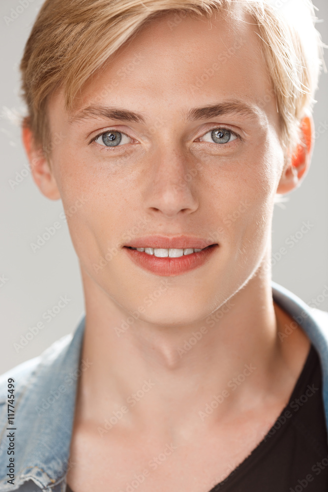 Close up portrait of handsome smiling blond young man wearing casual black t-shirt and jeans jacket looking in camera on grey background - obrazy, fototapety, plakaty 