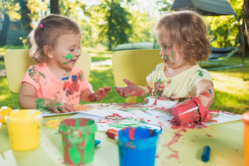 Two-year old girls painting with poster paintings together against green lawn