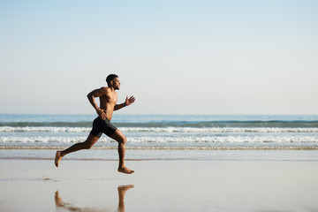 Black fit man running barefoot by the sea on the beach. Powerful runner training outdoor on summer.