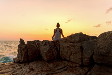 Wall Mural - Young woman meditating on the rocks by the sea on sunrise background