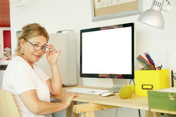 Beautiful mature blonde female pensioner in spectacles looking at camera while communicating with her grandchildren online, sitting in front of computer screen with copy space for your advertisement