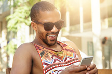 Young Attractive Dark-skinned Male Looking Happy Holding His Cellphone, Receiving Good News From Sister About Birth Of His Niece. African Man In Trendy Sunglasses Liking Funny Posts On Social Network