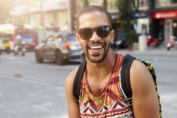 Outdoor portrait of fashionable dark-skinned traveller dressed like hipster, wearing backpack and stylish shades, walking down a busy street, looking happy and cheerful, having fun in a big city