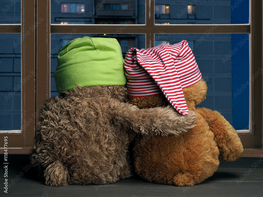 Two friends looking out the window at the windows of the house. Toys colorful hats bear cubs. Embrace the . Concept - love, friendship, support, evening together - obrazy, fototapety, plakaty 