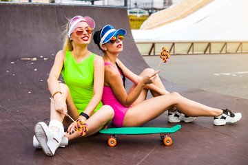 Wall Mural - Fashion glamor stylish swag young girl model blonde and brunette with perfect tanned bodies in bright sexy bathing suits in a skate park, Long board, lady laughing and having fun, licking lollipops