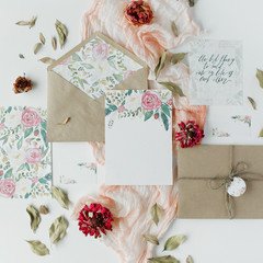 Workspace. Wedding invitation cards, craft envelopes, pink and red roses and green leaves on white background. Overhead view. Flat lay, top view