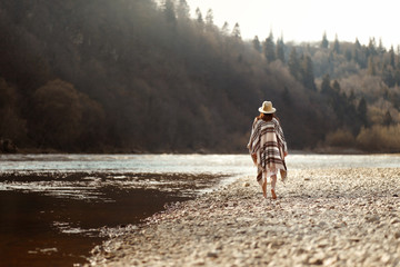 Canvas Print - beautiful woman hipster walking on river beach in mountains,wearing hat and poncho, boho travel concept, space for text