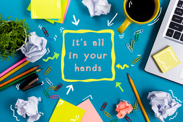 It is all in your hands. Office table desk with supplies, white blank note pad, cup, pen, pc, crumpled paper, flower on blue background. Top view