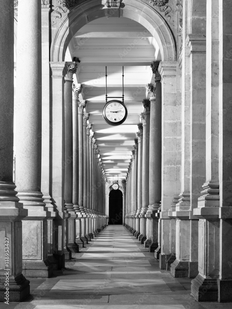 Long colonnafe corridor with columns and clock hanging from ceiling. Cloister perspective. . Black and white image. - obrazy, fototapety, plakaty 