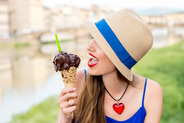 Wall Mural - Young female tourist in blue dress holding chocolate ice cream in waffle cone sitting near the river in Florence city. Eating traditional italian ice cream gelato.