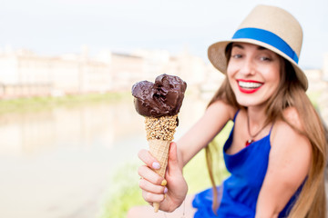 Wall Mural - Young female tourist in blue dress holding chocolate ice cream in waffle cone sitting near the river in Florence city. Image with small depth of field focused on ice cream