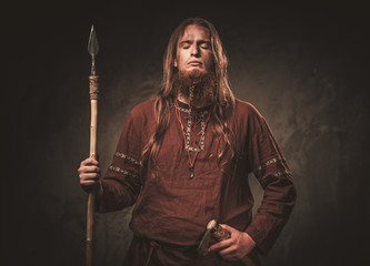 Serious viking with a spear in a traditional warrior clothes, posing on a dark background.