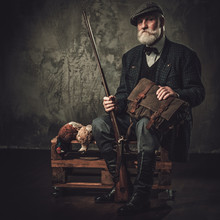 Senior Hunter With A Shotgun And Pheasants In A Traditional Shooting Clothing, Sitting On A Dark Background.
