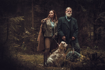 Two hunters with dogs and shotguns in a traditional shooting clothing, posing on a dark forest background.