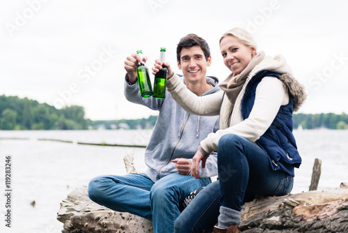 Fotografía couple sitting on tree stump at the riverside drinking beer