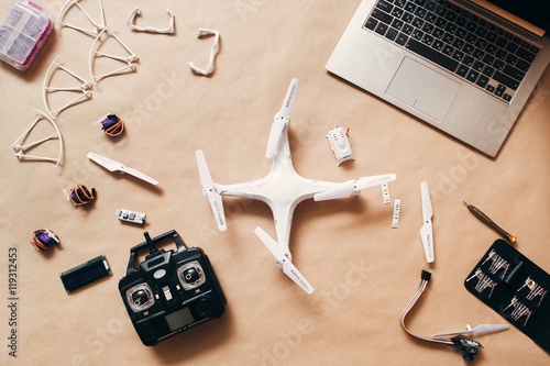 Plakat Dron z pilotem i laptopem, płaski lay. Widok z góry na stół z laptopem, biały quadrocopter z nadajnikiem i elementy do niego.