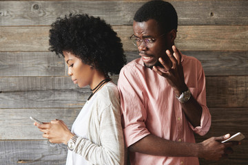 Portait of annoyed African man in glasses angry with his attractive girlfriend who is absorbed in surfing Internet and texting friends via social networks while standing back-to-back with her