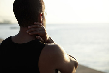 Rear view of muscular dark-skinned man in black A-shirt standing on embankment watching sun setting over horizon, thinking over problems in his life, rubbing his neck during evening walk. Flare sun