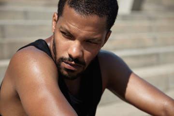 Wall Mural - Portrait of thoughtful dark-skinned male athlete having rest after intense training, sitting on pavement with serious and tired expression on his face against blurred background of gray concrete