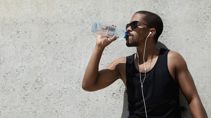 Wall Mural - Headshot of African jogger in sunglasses, drinking water, listen