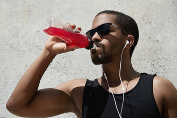 Wall Mural - Headshot of exhausted dark-skinned runner in shades and earphones wearing black sleeveless shirt, quenching his thirst with red juice or shake, relaxing after intense physical activity outdoors