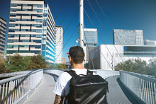 Hipster With A Large Black Backpack Travels On A Diverging Bridge Against City Landscape And Bright Blue Sky
