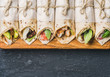 Tortilla wraps with various fillings on wooden board over dark grey grunge concrete background, top view, copy space, horizontal composition. Healthy snack or take-away lunch bites