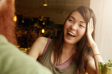 Selective focus. Good-looking girl with long dark hair laughing with mouth wide open, resting her elbow on table, having fun during first date with redhead man at restaurant with cozy interior