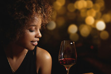 Profile Of Dark-skinned Luxurious Woman With Afro Haircut Wearing Elegant Evening Dress Drinking Red Wine, Sitting At Restaurant Against Blurred Background For Your Text Or Advertising Content