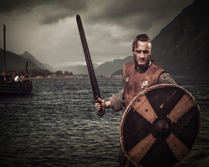 Wall Mural - Viking warrior in the attack, standing along the shore with Drakkar and mountains on the background.