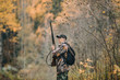 © scharfsinn86 - Male hunter in the woods. Man in camouflage with a backpack. Autumn forest, leaf fall.