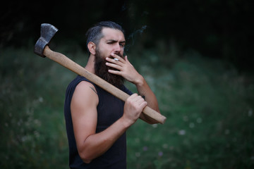 Wall Mural - Brutal brunette bearded man in warm hat with a hatchet in the woods on a background of trees