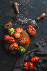 Wall Mural - Fresh colorful ripe Fall heirloom tomatoes on wooden board, herb chopper knife for cooking or salad making over grunge dark plywood background, top view. Harvest vegetable cooking conception.