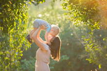 Happy Young Mother Playing With Her Littlte Baby Son On Sunshine Warm Autumn Or Summer Day. Beautiful Sunset Light In The Apple Garden Or In The Park. Happy Family Concept