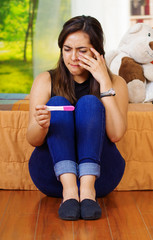 Wall Mural - Pretty young brunette woman sitting on floor holding pregnancy home test, crying wiping tears, looking emotional, garden window background