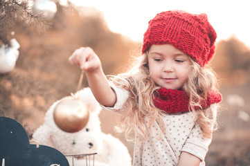 Wall Mural - Smiling kid girl 4-5 year old holding christmas ball wearing knitted red hat and scarf outdoors. Winter portrait. Christmas holidays.