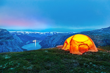 orange lighting tent in norwegian mountains against fjord background in night.